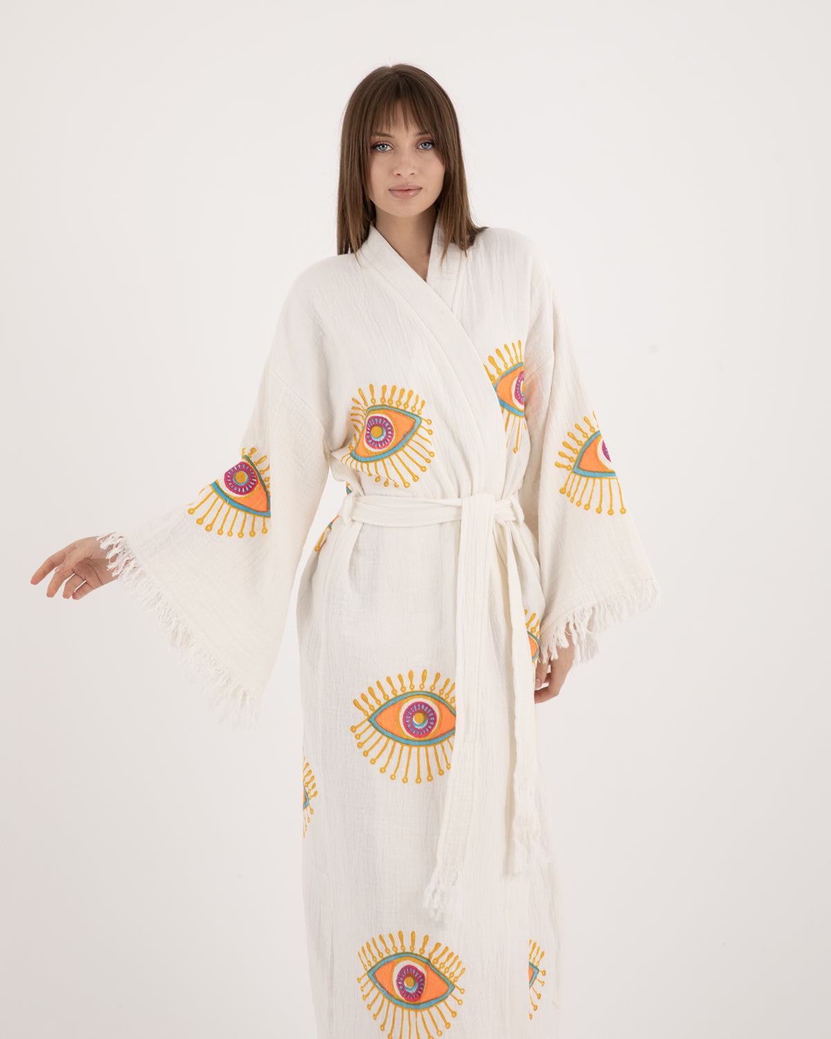 A woman modeling a long, lightweight white Turkish kimono-kaftan robe featuring vibrant evil eye motifs on the sleeves and body.