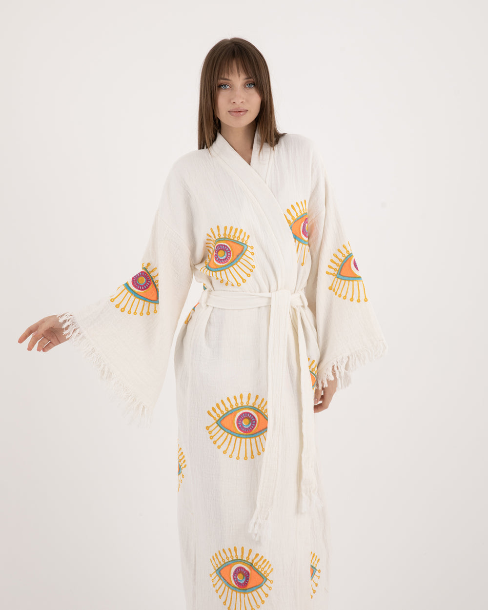 A woman modeling a long, lightweight white Turkish kimono-kaftan robe featuring vibrant evil eye motifs on the sleeves and body.