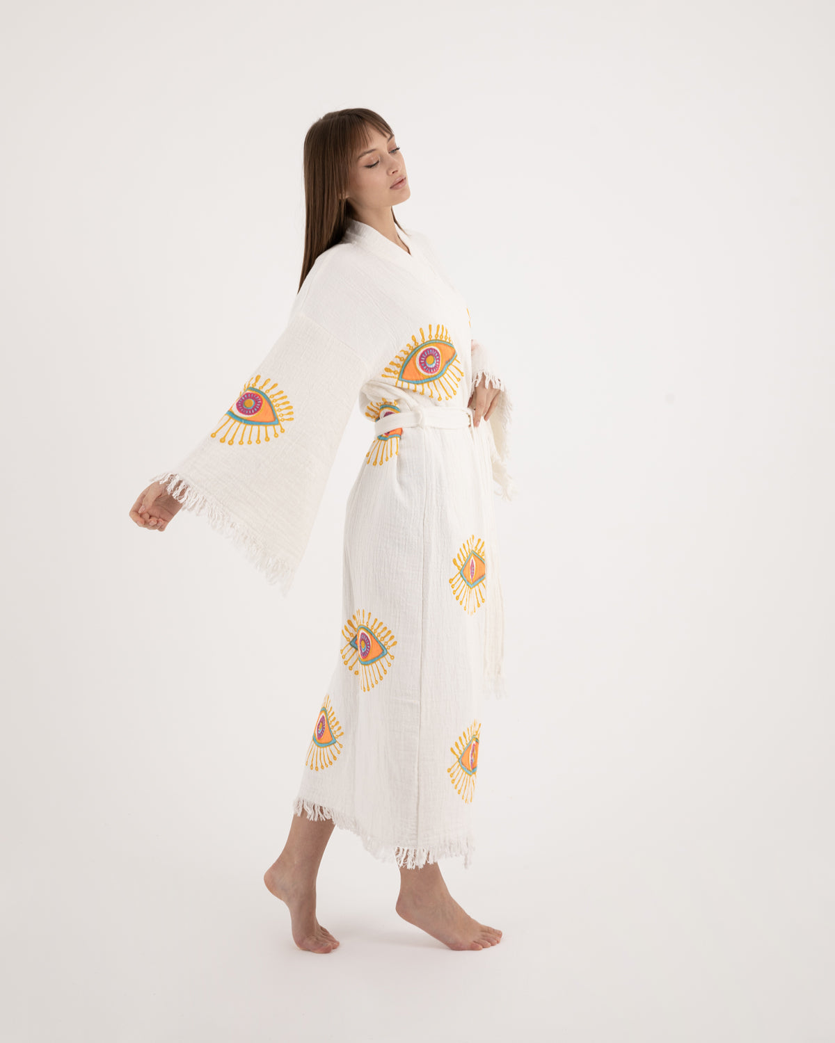 A woman modeling a long, lightweight white Turkish towel kimono robe featuring vibrant evil eye motifs on the sleeves and body.