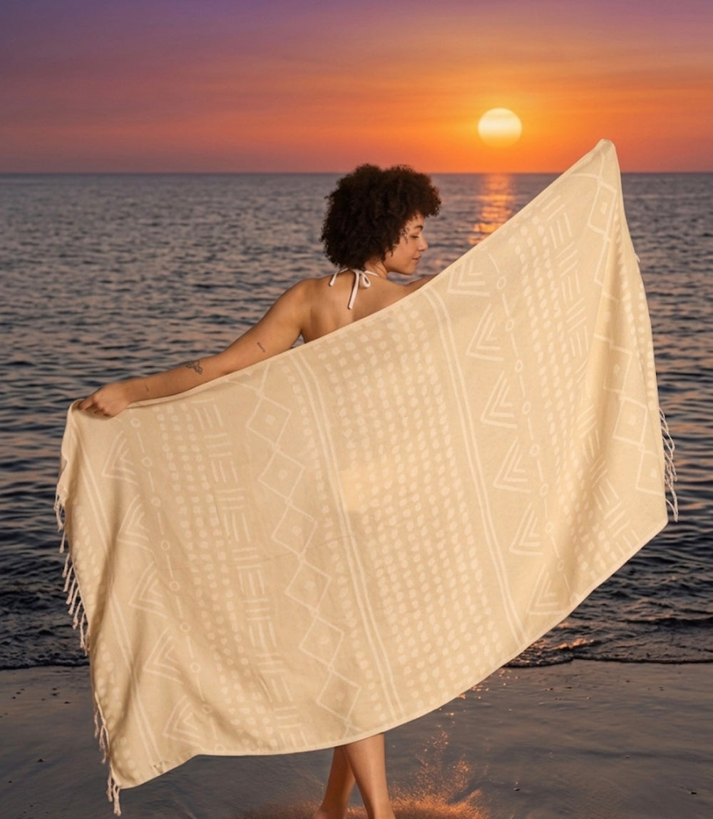 A woman by the sea at sunset holding up a lightweight, sand free cream-colored Turkish towel with traditional ethnic woven designs, showcasing its texture and drape. Made with organic Turkish cotton 