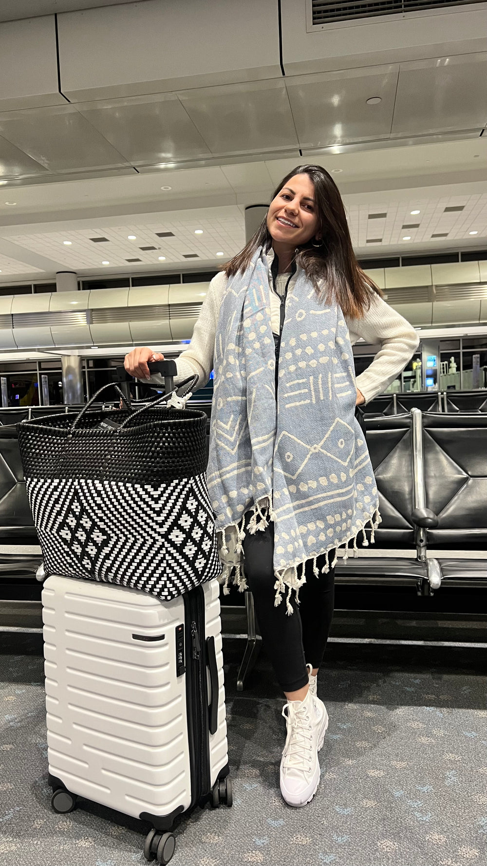 Woman wearing a light blue ethnic Turkish towel scarf with tassels posing in an airport terminal, showcasing trendy travel outfit accessories and organic cotton peshtemal style.⁠