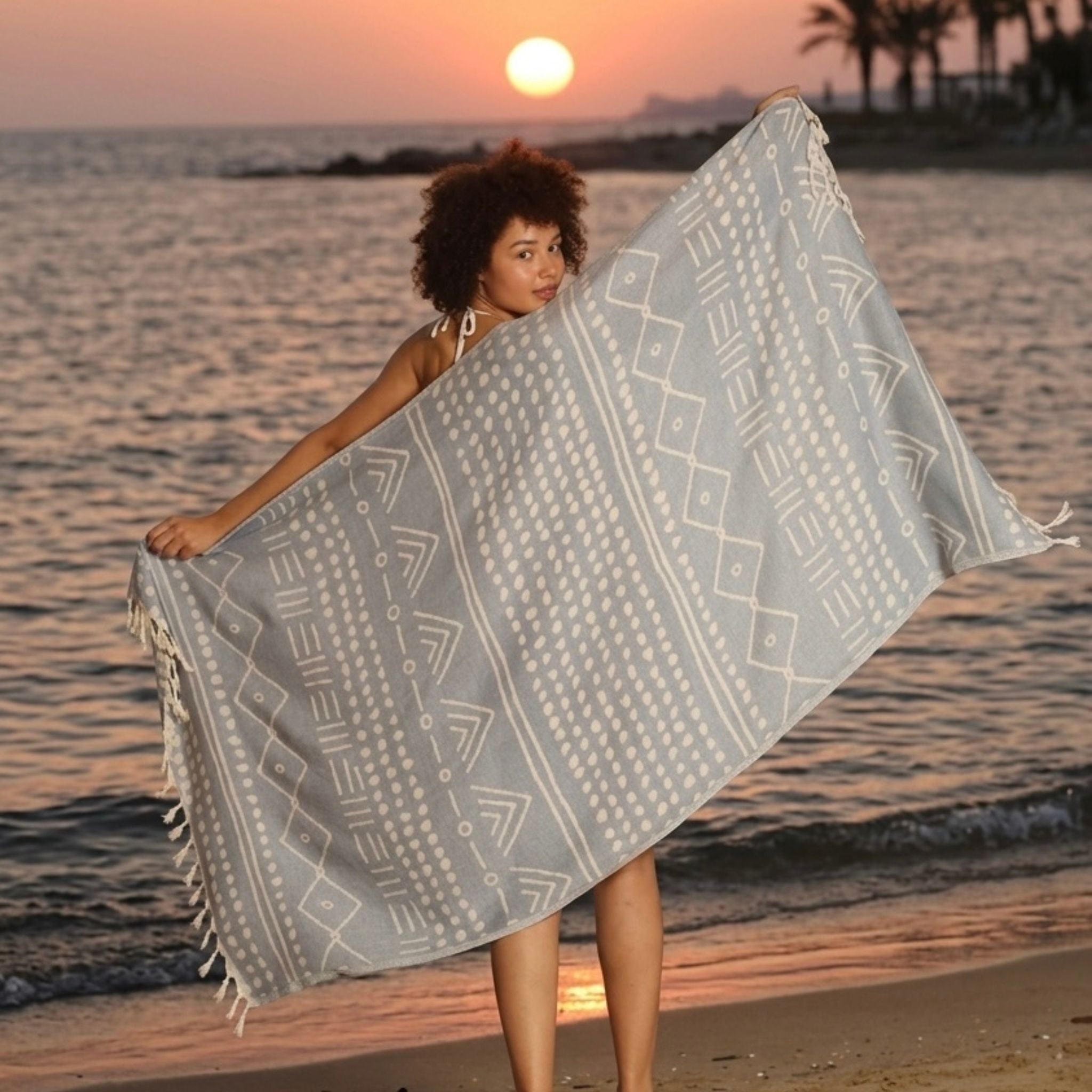 Lightweight blue Turkish cotton towel with ethnic tribal motifs and tassels displayed on a beach at sunset.