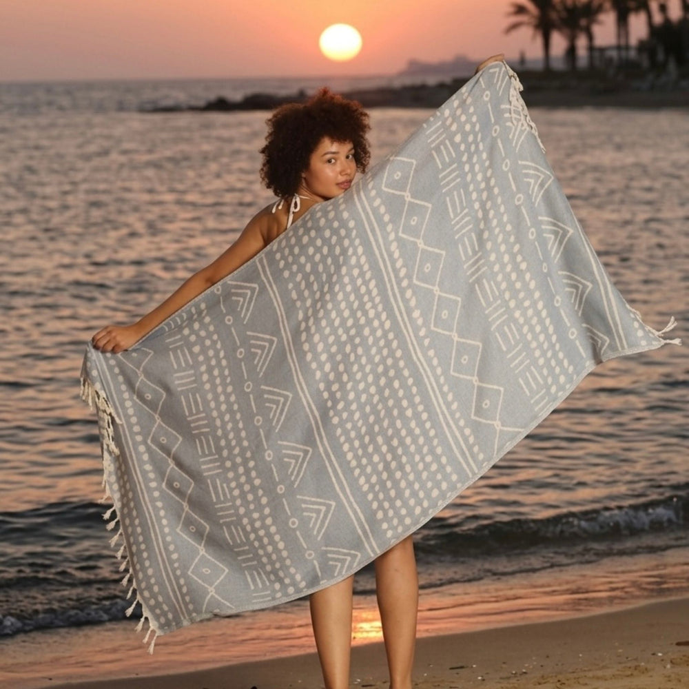 Lightweight blue Turkish cotton towel with ethnic tribal motifs and tassels displayed on a beach at sunset.